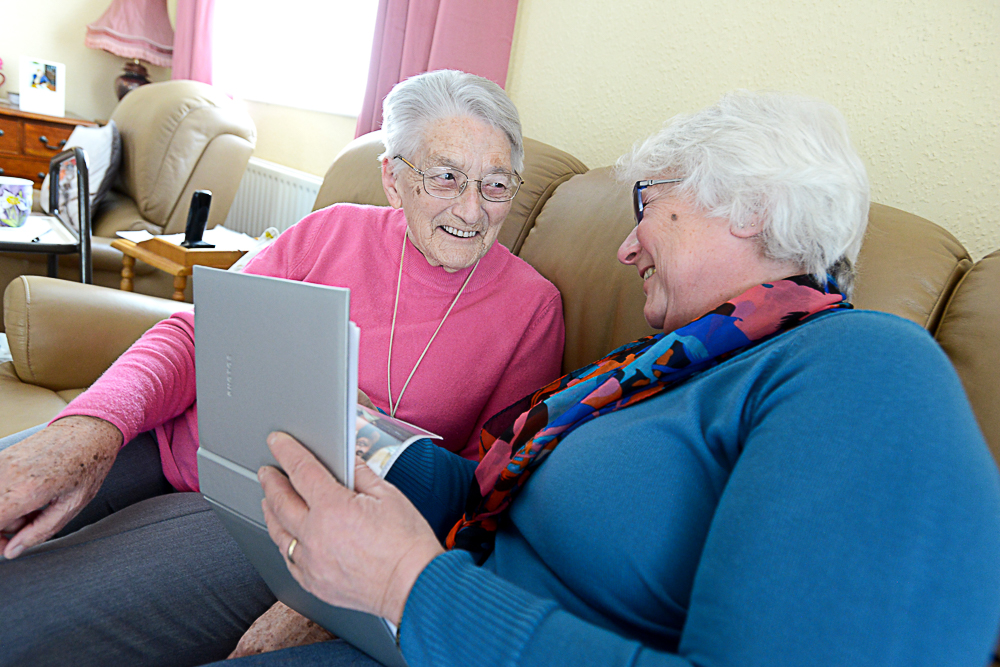 This photo 2 women sat on a sofa looking at an ipad and smiling at each other.